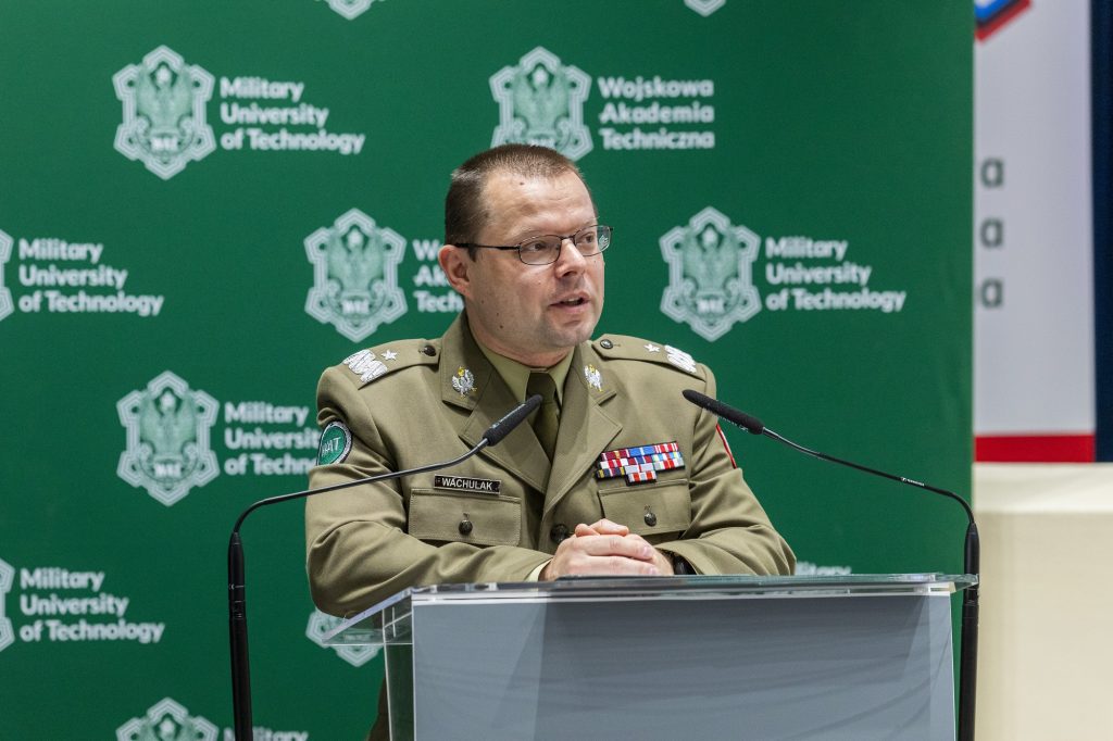 Generał brygady profesor doktor habilitowany inżynier Przemysław Wachulak, Rektor-Komendant WAT stojący za mównicą, wygłaszający przemówienia podczas uroczystego otwarcia konferencji, w tle ścianka z logotypem WAT